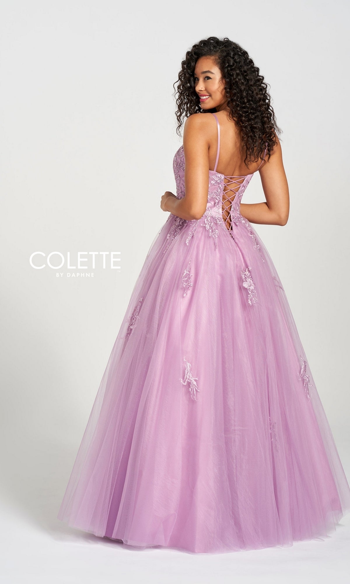 A woman faces away, wearing the Long Prom Dress CL12212 by Colette, a floor-length lavender tulle gown featuring a lace-up back, floral appliqué accents, and delicate lace embroidery for an elegant look.
